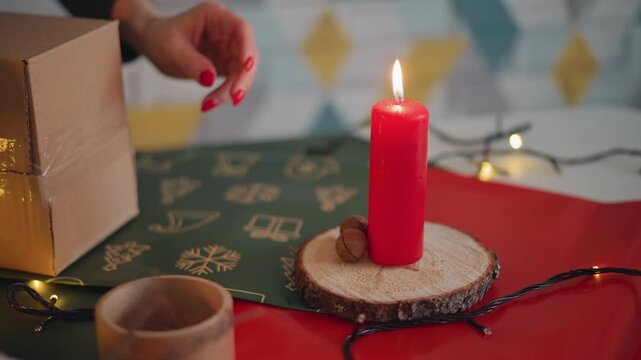 Caucasian hands crafting candle centerpiece near present box, string lights glowing, wood slab base with pinecone placement, red candle flame, festive wrapping materials and tape, cozy joyful holiday