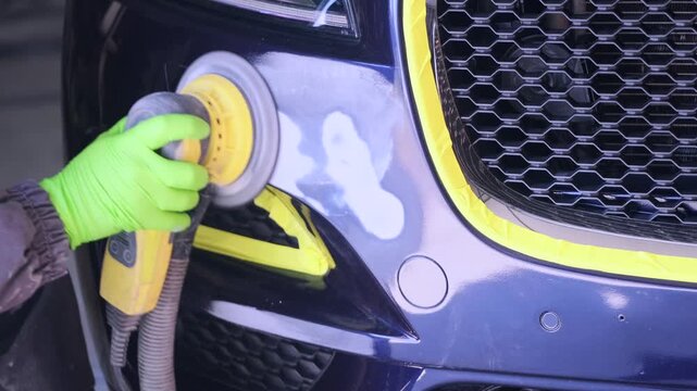 Auto technician polishing damaged car bumper close-up with orbital sander in repair garage, preparing vehicle surface for urgent local repainting service.