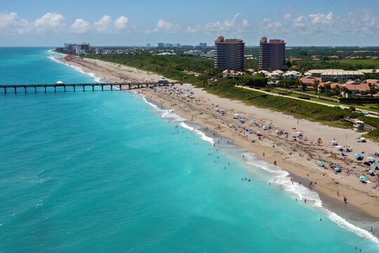 Aerial view of Juno Beach Pier stretching into the turquoise Atlantic Ocean with people on the sandy shore and coastal buildings Juno Beach, Florida, United States.