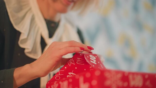 Closeup caucasian woman wrapping red present in boutiquestyle interior with patterned wallpaper and ruffled blouse, neat folds and ribbon, smiling as she prepares elegant gift with playful holiday