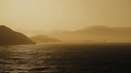 Fototapeta premium Coastal mountains slope into a shimmering ocean under warm, hazy light as distant peaks fade into soft atmospheric glow at golden hour, calm and serene