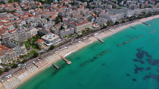 4K aerial view of Boulevard de la Croisette in Cannes with private beach clubs, umbrellas in rows, belle epoque palace hotels, wooden jetties and crystal turquoise Mediterranean sea.