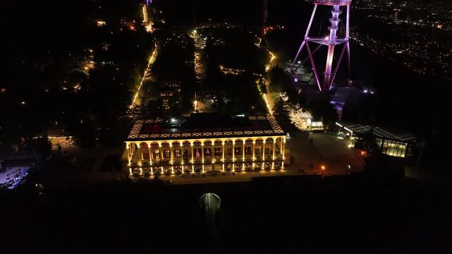 Aerial night view of Mtatsminda Park restaurant and funicular station in Tbilisi, Georgia, featuring illuminated arched architecture