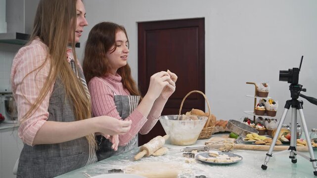 Mother and teen daughter filming bakery vlog with camera on tripod in kitchen. Family food bloggers recording video for social media channel, domestic lifestyle, homemade cookies cooking tutorial