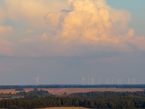 Aerial view of wind turbines standing across a rural landscape with agricultural fields and forests under massive cumulus clouds at sunset Taujenai, Vilnius County, Lithuania.