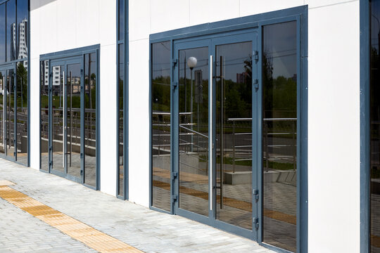 Dark gray aluminum double doors with large glass panels installed in white building facade. Modern architectural design features long vertical handles and matching side windows along paved sidewalk.