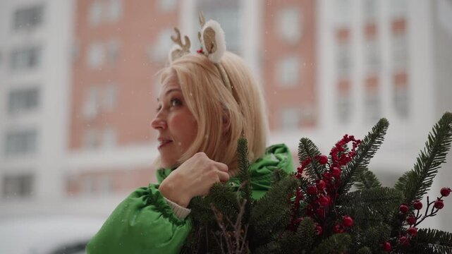 White woman reindeer headband holding bouquet, playful holiday shopper adjusting antler ears, snowy urban backdrop with modern buildings, green coat and red berries contrast, laughing glance