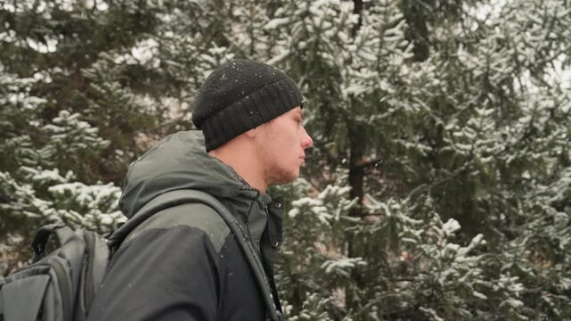 Man commuter walking snowy city park, purposeful profile with backpack and beanie, flurries drifting past residential buildings and evergreen trees, side view captures breath in cold air and steady