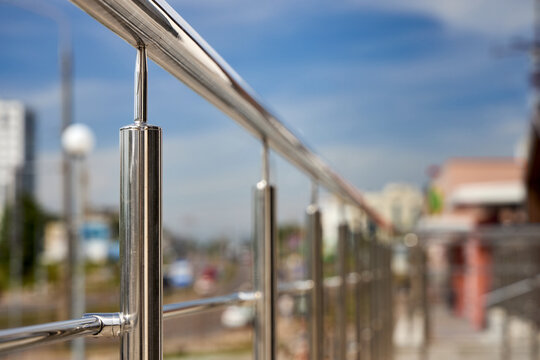 Polished stainless steel railing stands against blurred urban backdrop under blue sky. Modern metallic structure features sleek handrails and vertical balusters reflecting bright daylight.