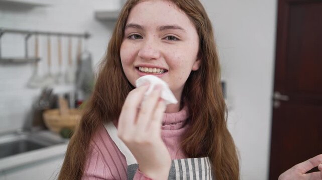 ็Happy teenage girl with freckles smiling and wiping her face with a white napkin in kitchen. Cute young baker during cooking class at home, domestic lifestyle