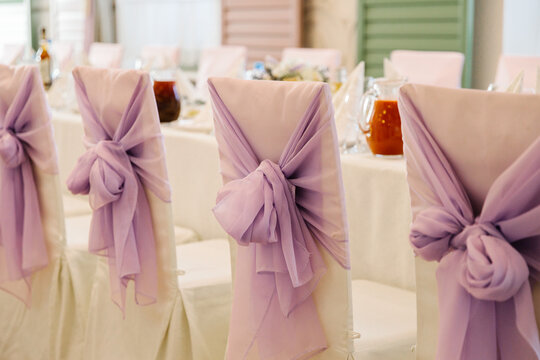 Elegant chairs decorated with purple sashes and bows are set for a formal event or wedding reception, with a table featuring drinks in the background, creating a festive atmosphere.