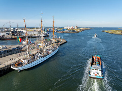 Velsen, Netherlands - 19 August 2025: Aerial view of a large tall ship, the Portuguese NRP Sagres, docked at the pier and a barge navigating the harbor waters under a clear blue sky.