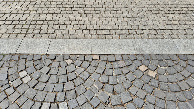 A high-angle shot of two cobblestone patterns separated by a granite border: light beige stones in straight rows above, and dark gray stones in a decorative fan-shaped arrangement below.