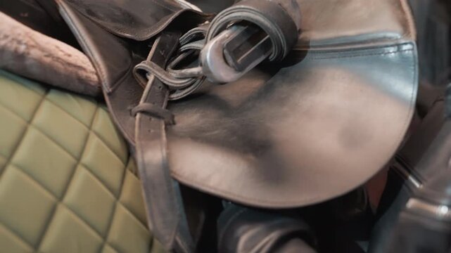 Caucasian trainer tightening leather saddle strap in busy indoor barn with olive quilted pad visible, closeup on gloved hands securing stirrup and buckle, urgent lastminute check, focused expression,