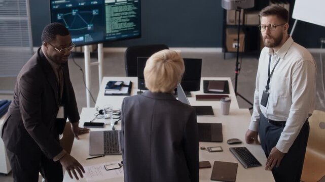 High angle zoom out portrait of three diverse cybersecurity engineers having discussion in control center and looking at camera
