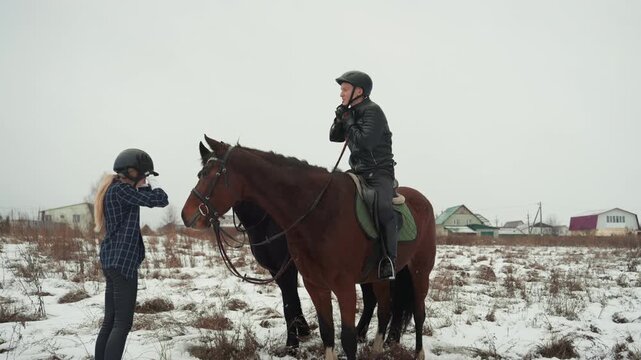 Caucasian woman instructing mounted caucasian man snowy field lesson focusing reins and balance helmeted rider listens coach gestures supportive touch visible winter breath distant village houses calm