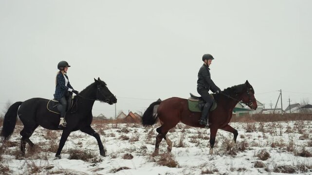 Two white riders on horses trotting snowy field, instructor guiding student during tandem riding lesson, helmets and saddles visible, rustic village backdrop, cold overcast sky, focused training,