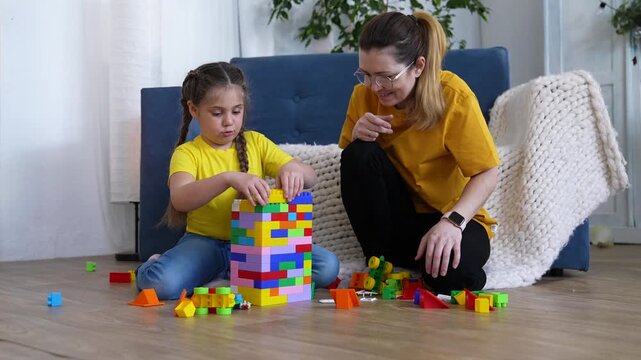 Mother and daughter play together on floor. Happy family build tall tower. Mother helps child with colorful toy block. Young daughter gently stacks plastic toy block. They play inside quiet home.