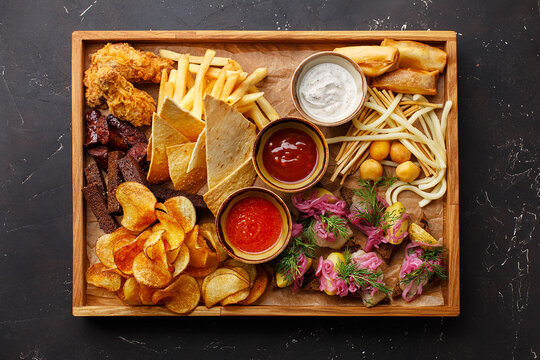 Set of assorted beer snacks and sauces served in a wooden box on dark concrete background. Potato chips, nachos, french fries, rye bread croutons, sausages, spring rolls, cheese, herring toasts