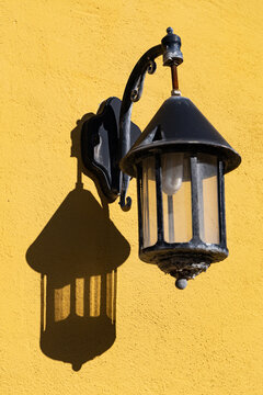 View of a vintage black outdoor lantern casting a sharp shadow on a bright yellow textured wall in Ribe, Denmark.