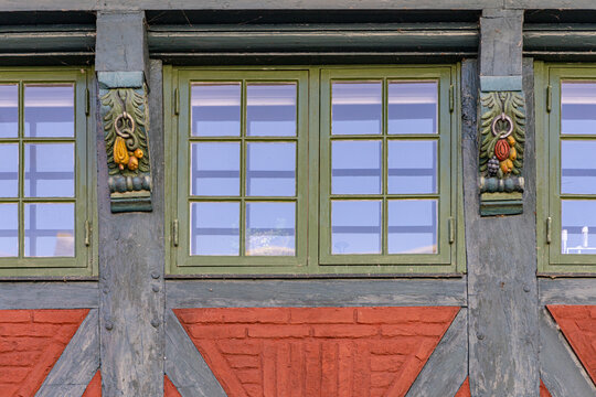 View of traditional half-timbered house facade featuring green windows, ornate carved wooden corbels with fruit motifs, and red brickwork in Ribe, Denmark.