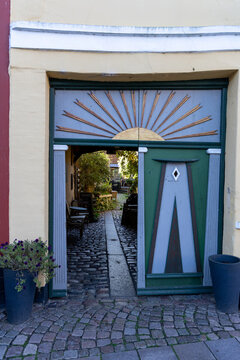 View of a decorative green wooden gate with a sunburst motif leading to a cobblestone courtyard with outdoor seating and lush greenery in Ribe, Denmark.