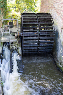 View of a large historic water wheel rotating with rushing water next to a traditional brick wall and green foliage in Ribe, Denmark.