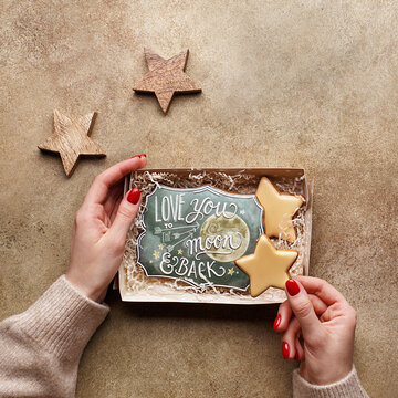 Female hands in sweater holding cardboard gift box with decorated gingerbread cookies in shape of stars and with "Love you to the moon and back" text over beige concrete background. Overhead view