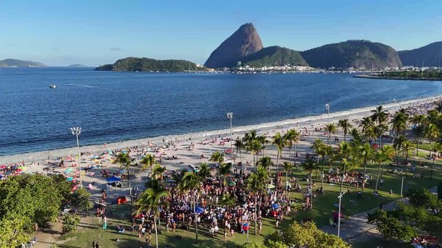 Aerial drone footage showing a large crowd gathered for a carnival rehearsal near Parque do Flamengo with Sugarloaf Mountain and Guanabara Bay