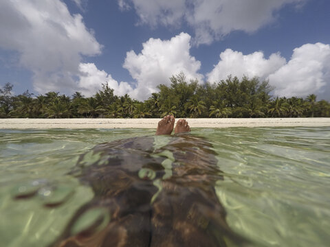 View of Tiwi Beach with a person's feet floating in clear turquoise water near the white sand shore and palm trees Tiwi, Wilaya ya Kwale, Kenya.