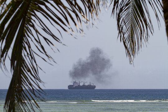 View of a large cargo ship emitting dark smoke on the horizon framed by tropical palm fronds at Tiwi Beach Tiwi, Wilaya ya Kwale, Kenya.