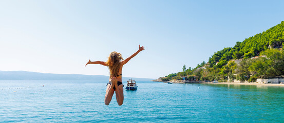 Happy young woman in bikini jumping into turquoise sea water with arms raised during summer vacation © M.studio