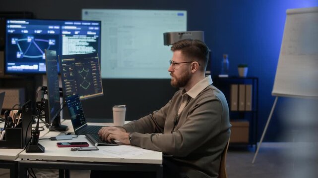 Side view of focused Caucasian analyst typing on computer keyboard while sitting at table working in network center