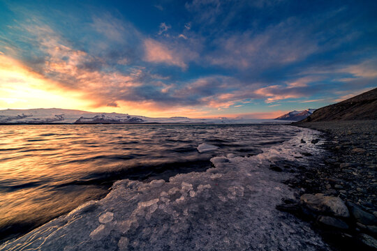 View of Jokulsarlon Glacier Lagoon with floating ice chunks and distant snow-capped mountains under a vibrant sunset sky Sveitarfelagid Hornafjordur, Iceland.