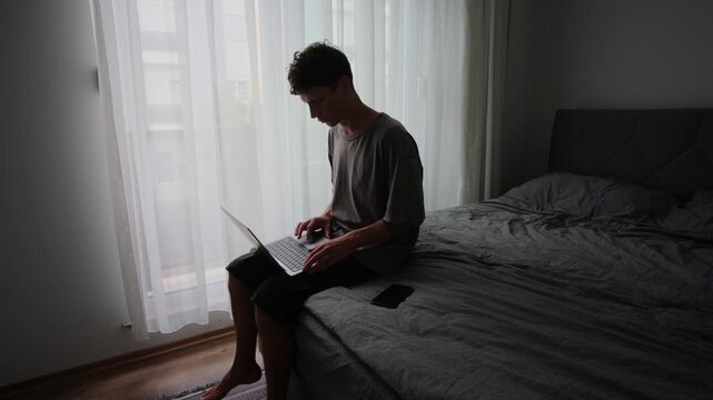 White man working laptop by window, soft backlit curtains, seated on bed edge, contemplative silhouette, minimal bedroom, slow morning routine, focused remote