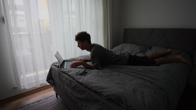 White man lying on bed laptop, prone position, designer coding with tense posture, natural light through curtains, informal duvet setup, late night creative