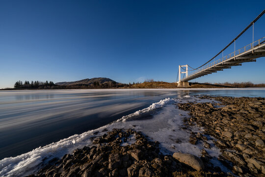 View of Laugaras Bridge spanning a frozen river with ice floes and rocky banks under a clear blue sky in Blaskogabyggd, Iceland.