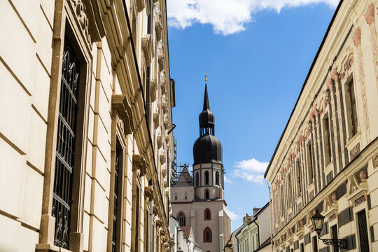 View of the historic St. Nicholas Basilica tower framed by ornate classical facades along a narrow cobblestone street under a bright blue sky Trnava, Trnava Region, Slovakia.