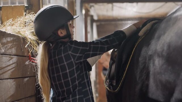 Helmeted rider fitting saddle onto horse in barn aisle, tightening girth and checking balance with careful hands bright stable light, hay bales, concentrated instruction and teamwork atmosphere