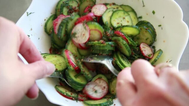 Close-up of preparing a simple summer salad by tossing sliced cucumbers and radishes with mustard dressing in a large salad bowl