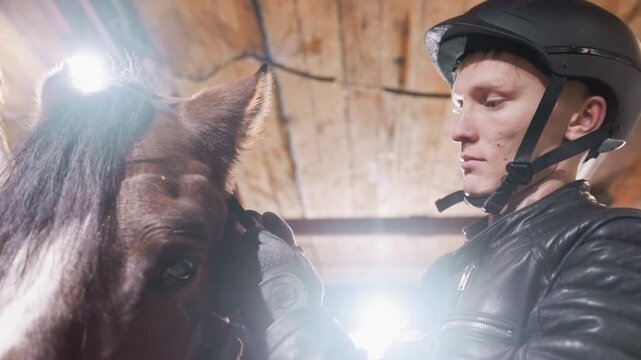Closeup white trainer adjusts horse bridle inside rustic stable, helmeted man concentrates while checking tack and grooming, warm overhead light, leather jacket hands steady, intimate equine care
