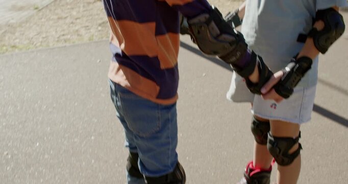 Children hold hands practicing standing on inline rollerblades on park pavement. Sister keeps balance steady while younger brother learns basic skate control safely