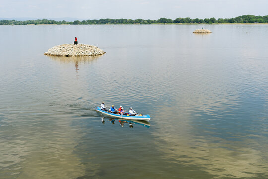 View of people paddling a blue canoe on calm water past small stone islands and a distant green shoreline in Bratislava, Bratislava Region, Slovakia.