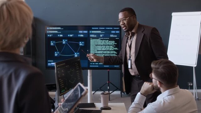 Medium long shot of young Black tech specialist presenting network data on screen to cybersecurity team while meeting in office