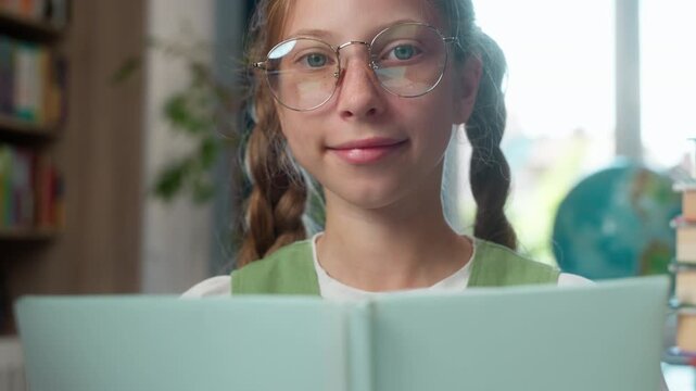 Happy clever Caucasian little girl child school kid in eyeglasses reading book turning pages looking at camera daughter smiling elementary studying in library pupil wearing glasses read study smile