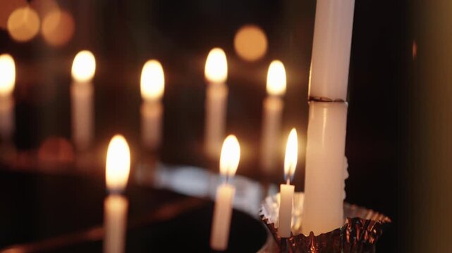 Multiple white candles burning on metal rack in dark shrine interior with warm bokeh background