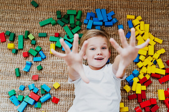 A smiling young boy relaxes on the floor among scattered bright wooden building blocks in a nursery. Authentic domestic life and creative heuristic play development concept.