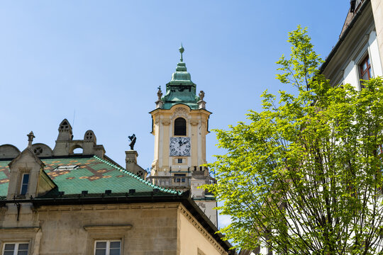 View of the historic Old Town Hall clock tower with its green copper roof and a vibrant green tree under a clear blue sky in Bratislava, Bratislava Region, Slovakia.