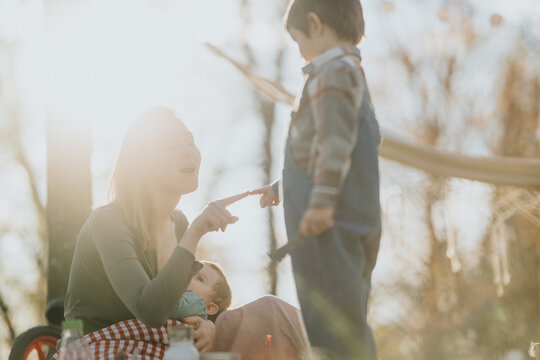 A mother sits outdoors holding a baby while interacting with her older child. Warm backlit scene shows family bonding during a casual outdoor moment.