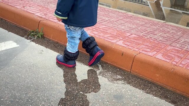 A little boy walks through puddles in rubber boots after the rain. 4k, slow motion.
